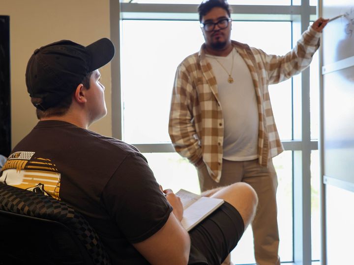 Two people talking across a room, one seated with a clipboard on his knee.